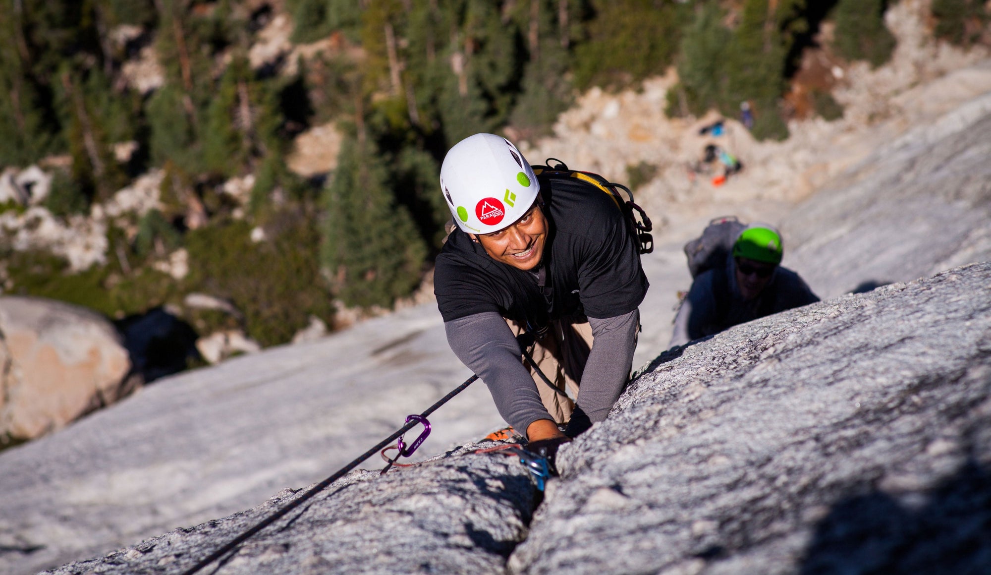 A climber smiling as the are half way up at rockwall with their climbing group behind them.