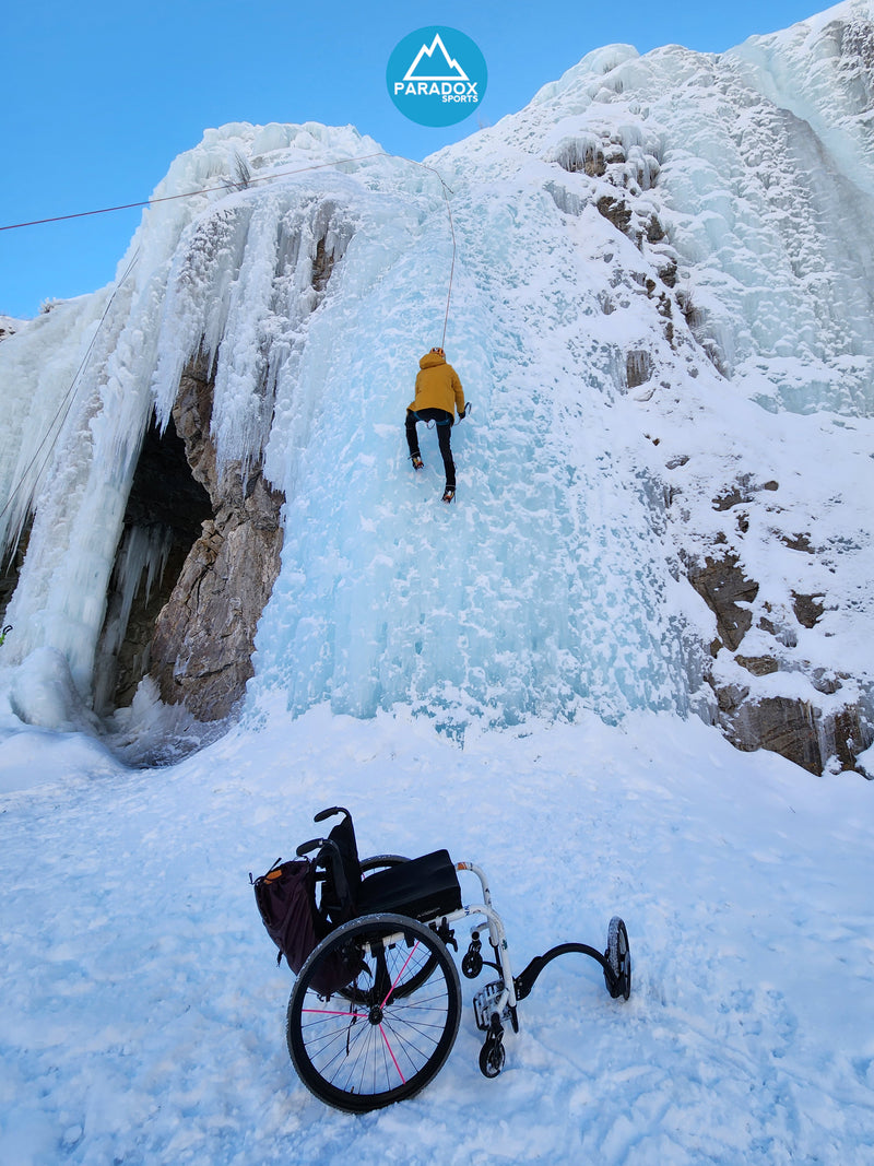 A climber halfway up an icewall with their wheelchair at the base.