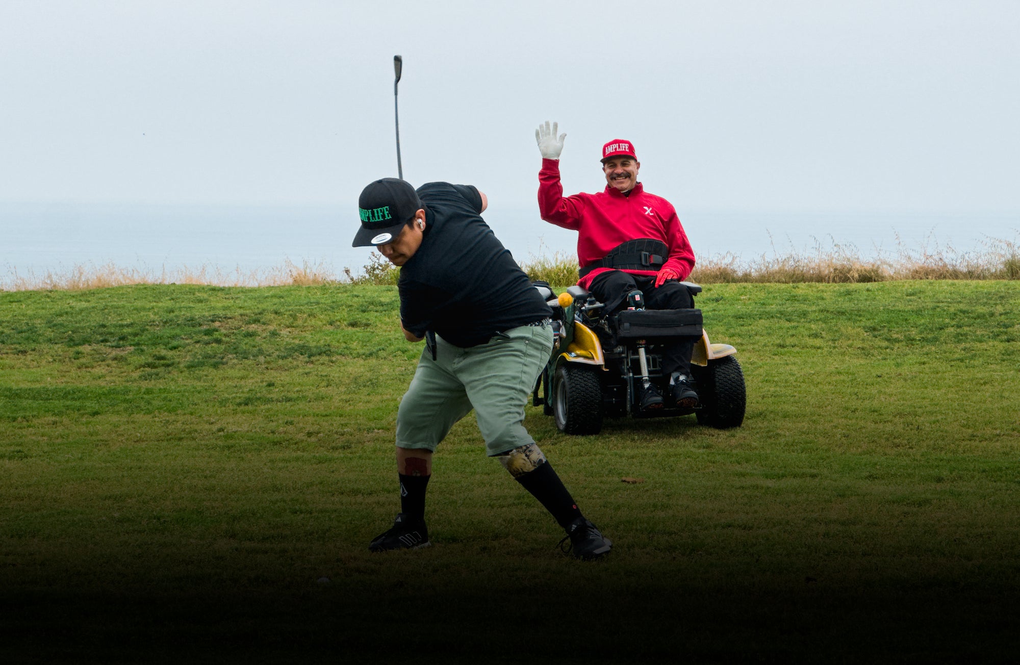 Amplife® Founder Abdul Nevarez wearing the Amplife® Flat Bill Snapback while smiling and putting his hand up behind Team Amplife® Ambassador Oscar Loreto, Jr. in his golf backswing while Oscar is wearing the Amplife® Curved Bill Snapback at Torrey Pines North 16th fairway.